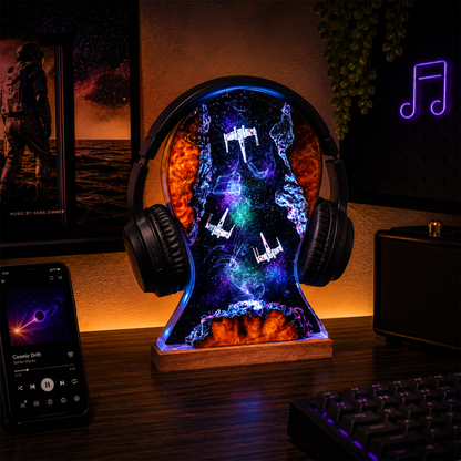 A headphone-shaped LED night light on a wooden base, glowing with a colorful galaxy and T-arkin space design, displayed on a desk with a smartphone, keyboard, and speaker in a room setting.