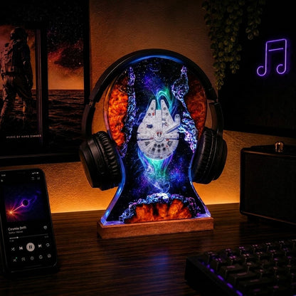 A decorative desk lamp shaped like a pair of headphones with a Millennium Falcon clock face inside, illuminated with multicolor LED lights on a wooden base. The lamp is shown on a dark wooden desk alongside a smartphone, keyboard, and speaker in a dimly lit room.