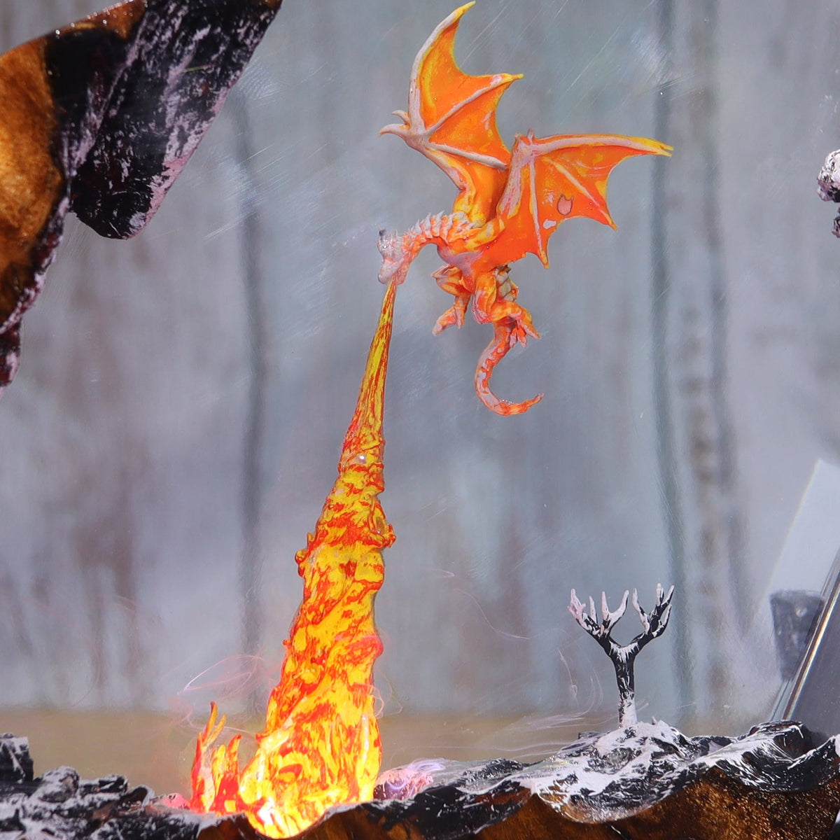 A fantasy dragon figurine displayed inside a clear acrylic display case with a glowing orange fire pit and miniature stone landscape, set against a rustic gray wooden background.