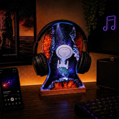 A headphone-shaped LED night light on a wooden desk, featuring a colorful cosmic space design with a white spaceship graphic and glowing blue, purple, and orange nebula effects. The light is shown in a styled home office environment with a smartphone, keyboard, and speaker nearby.