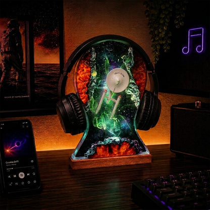 A headphone-shaped LED night light on a wooden desk, featuring a colorful space design with a white spaceship and glowing green nebula effects. The desk setup includes a smartphone, keyboard, and speaker in a dimly lit room.