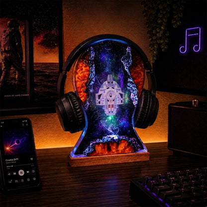 A decorative headphone-shaped LED night light on a wooden desk, featuring a colorful space design with a spaceship and glowing nebula effects. The lamp is styled in a gaming setup with a smartphone, keyboard, and speakers around it.