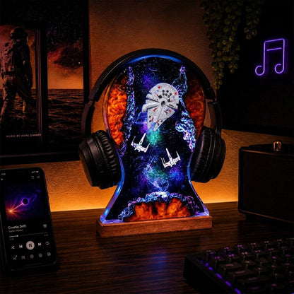 A headphone-shaped LED night light on a wooden desk, featuring a colorful space design with the Millennium Falcon and TIE fighters, flanked by black over-ear headphones. The scene is set in a dimly lit home office with a smartphone, keyboard, and speaker visible.